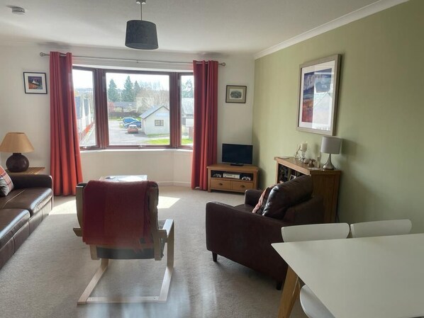 Ein Wohnzimmer mit einer Couch, einem Esstisch, einem Fernseher und einem Fenster mit Vorhängen.