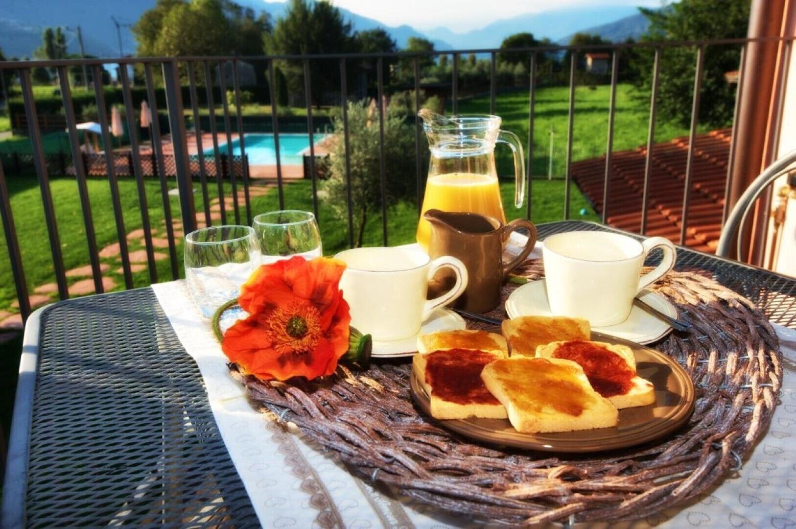 A table with breakfast items, a flower, and a view of a pool and mountains.