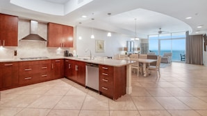 A modern kitchen with a large island, wooden cabinets, and a dining area with a view of the ocean. - Lazy River! D tower 1308D sleeps10 gulf front. (Orange Beach)