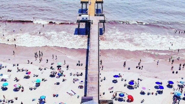 Une plage avec une jetée en bois qui s’avance dans l’océan, entourée de vacanciers et de parasols colorés.