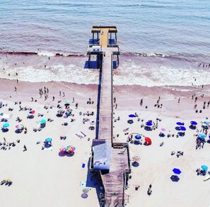 A beach with a wooden pier extending into the ocean, surrounded by beachgoers and colorful umbrellas. - Beachside! Views! Beautiful updated & steps to Beach & Boardwalk! (Ocean City)