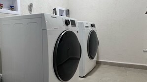 A laundry room with two white front-loading washing machines and a shelf with various items. - Seavetri Two - Modern apartment near serene beach and town centre (Speightstown)