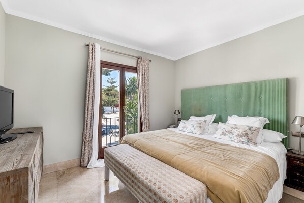 A bedroom with a bed, a TV, a bench, a green headboard, and a balcony with curtains.
