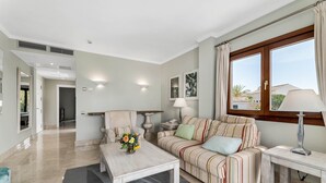 Een moderne woonkamer met een bank, fauteuil en salontafel. Er is een raam met gordijnen, een lamp en kunst aan de muur.