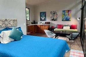 Une chambre avec un grand lit, un petit bureau et une chaise. On aperçoit un coin cuisine au fond de la pièce.