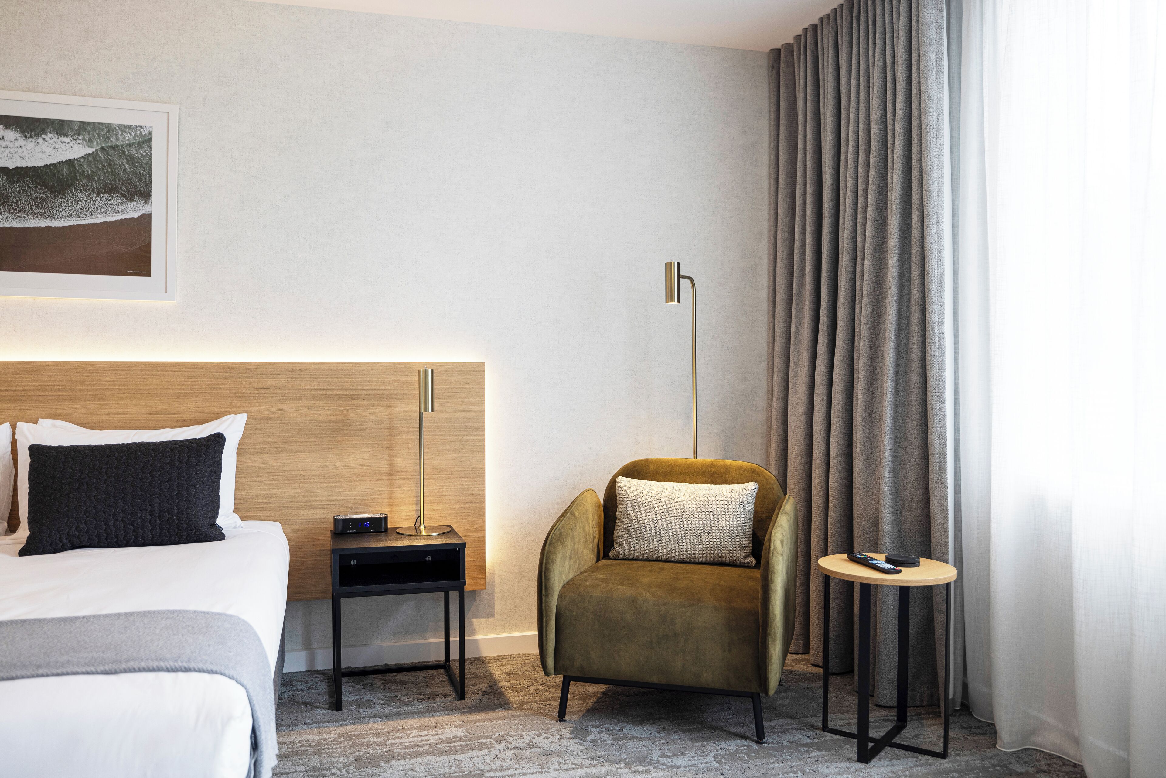 Una habitación de hotel con cama, mesita de noche, una silla, una pequeña mesa, una lámpara de pie y un ventanal grande con cortinas.