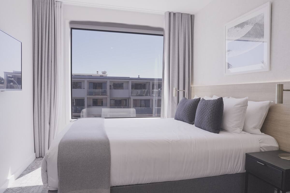 A hotel room with a large bed, a bedside table, a lamp, and a view of a building through the window.