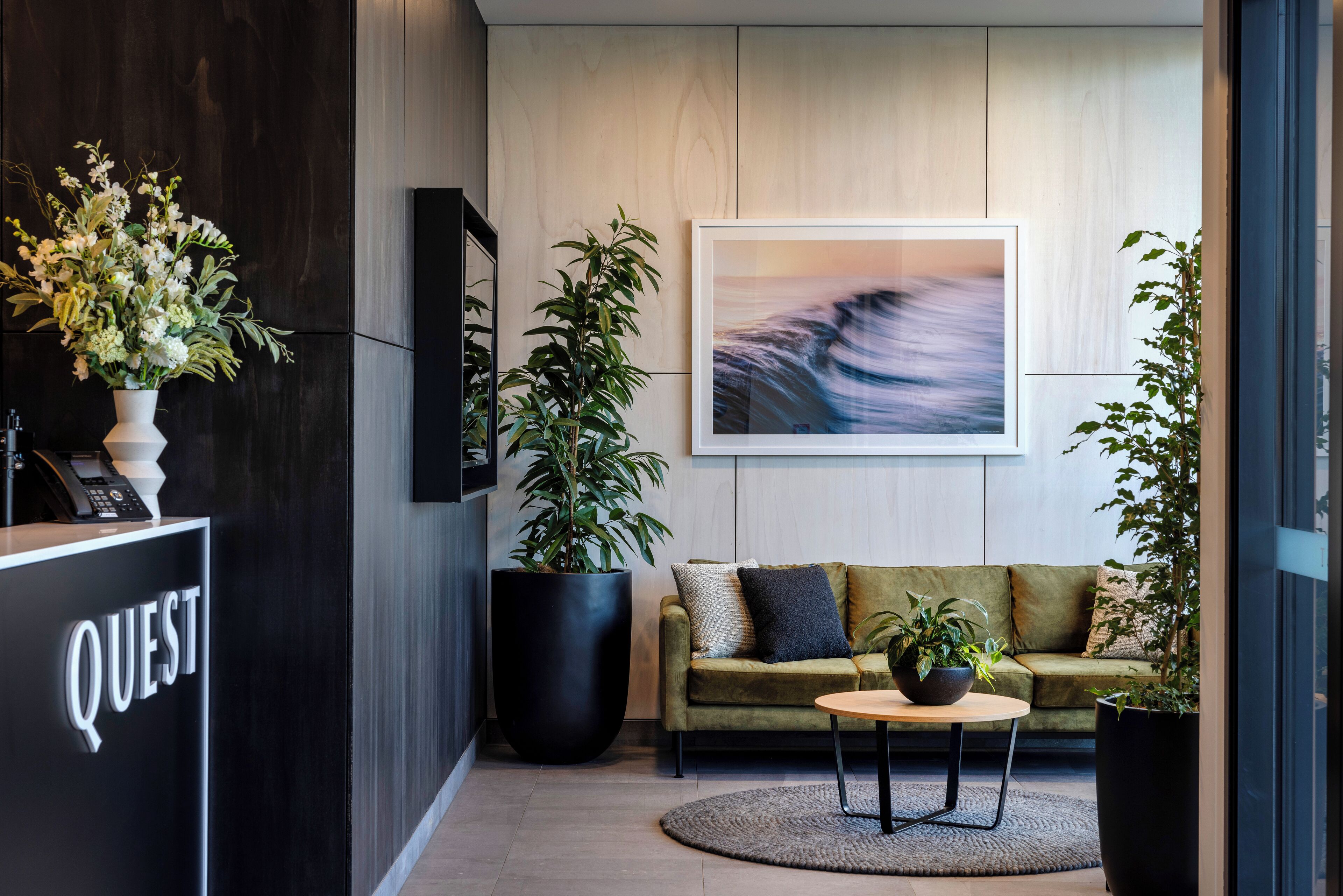 A modern lobby with a reception desk labeled "QUEST," a large potted plant, a green sofa, and a round table with a small plant.