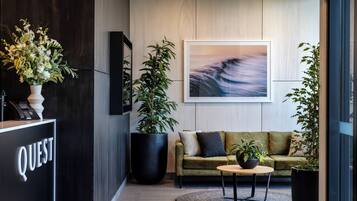 A modern lobby with a reception desk labeled "QUEST," a large potted plant, a green sofa, and a round table with a small plant.