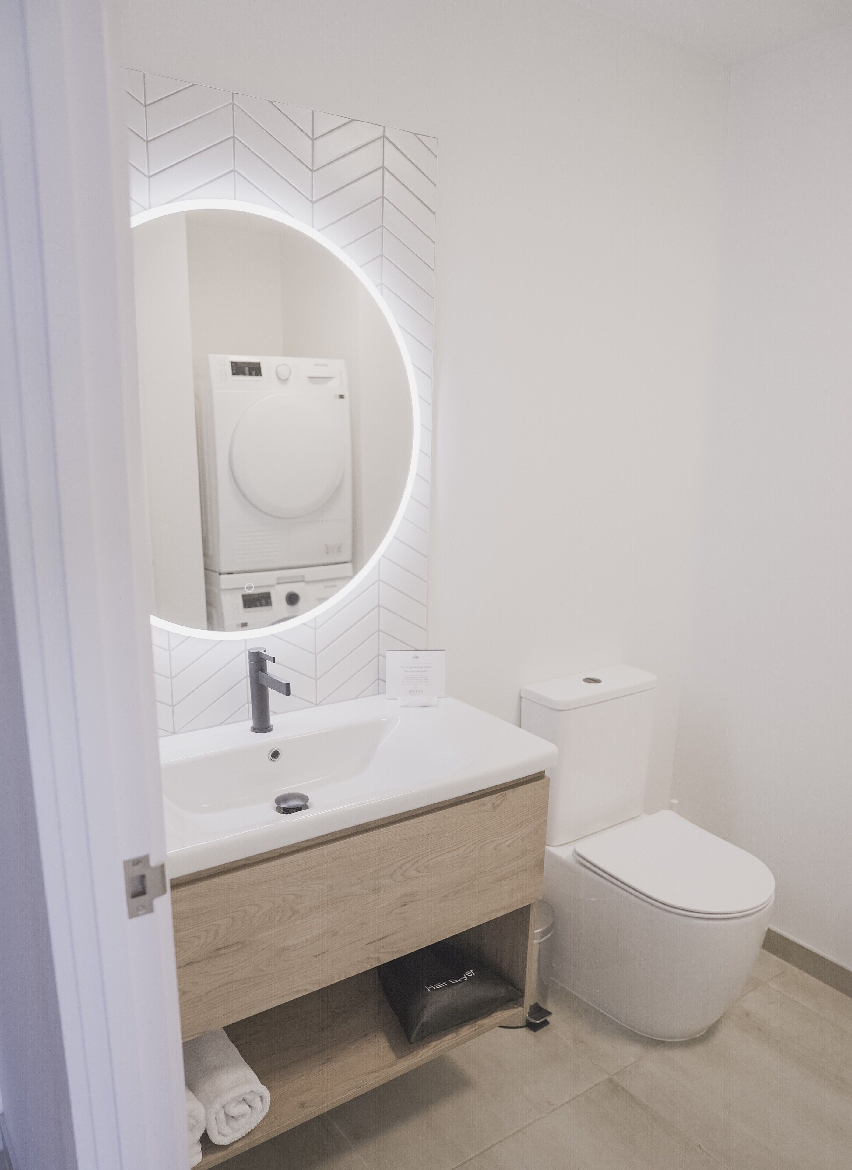 A modern bathroom with a white sink, a mirror with a circular frame, a toilet, and a towel rack.