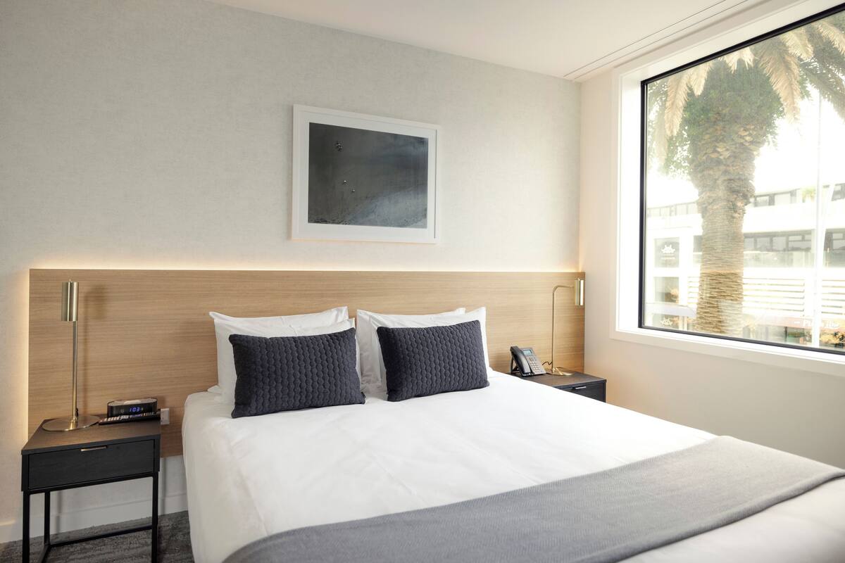 A modern hotel room with a large bed, two bedside tables, a nightstand, a framed picture on the wall, and a window with a view of palm trees.
