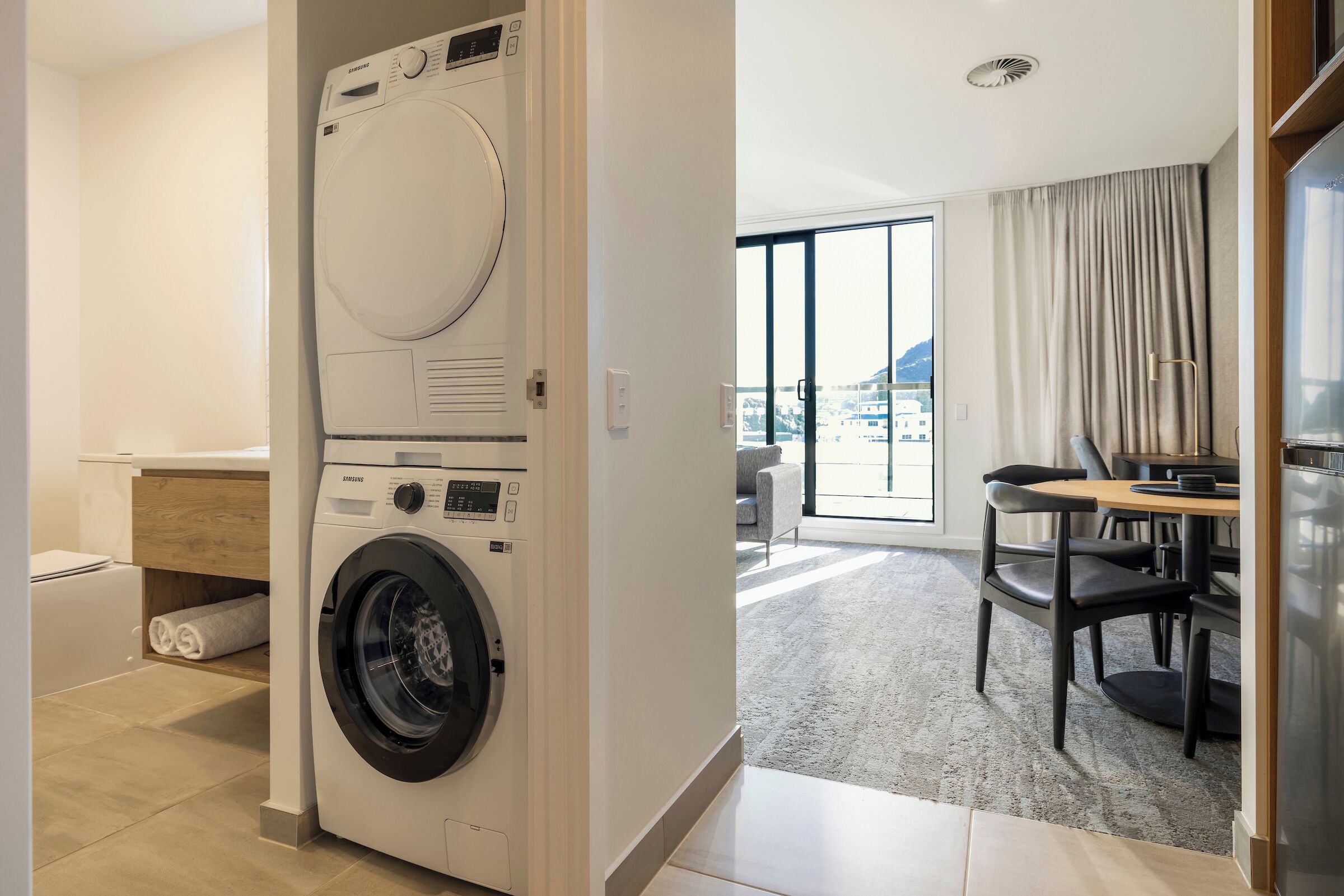 Een moderne hotelkamer met een ingebouwde wasmachine en droger, een eethoek met uitzicht en een groot raam met gordijnen.