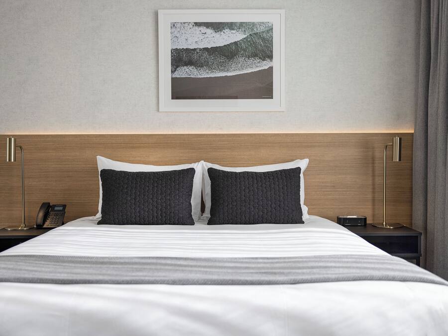 A neatly made bed with two pillows, a framed artwork above it, and a bedside table with a lamp and a phone.