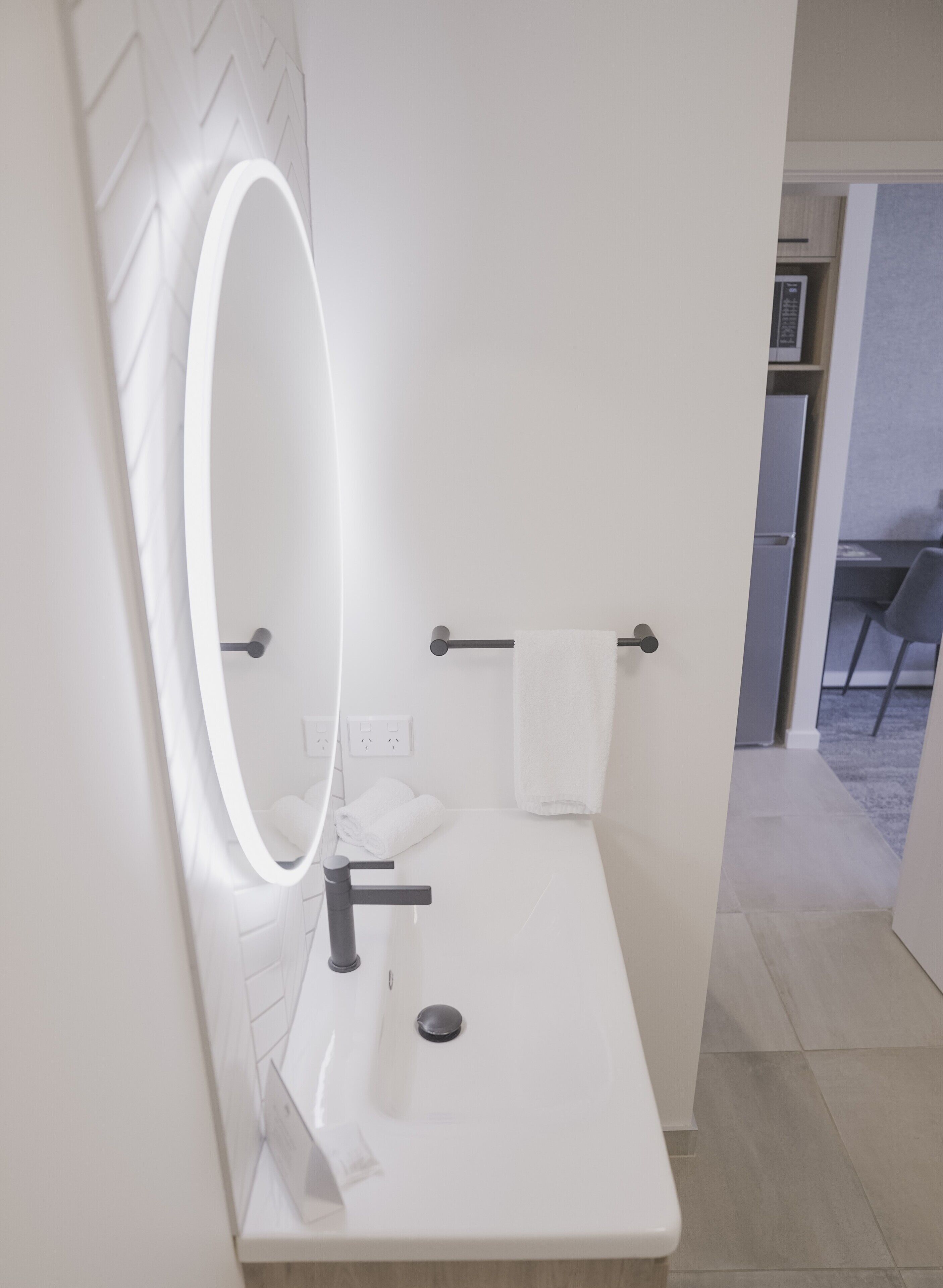 Een moderne badkamer met een witte wastafel, een spiegel met ingebouwde verlichting, een handdoekhouder en een zichtbare stopcontact.