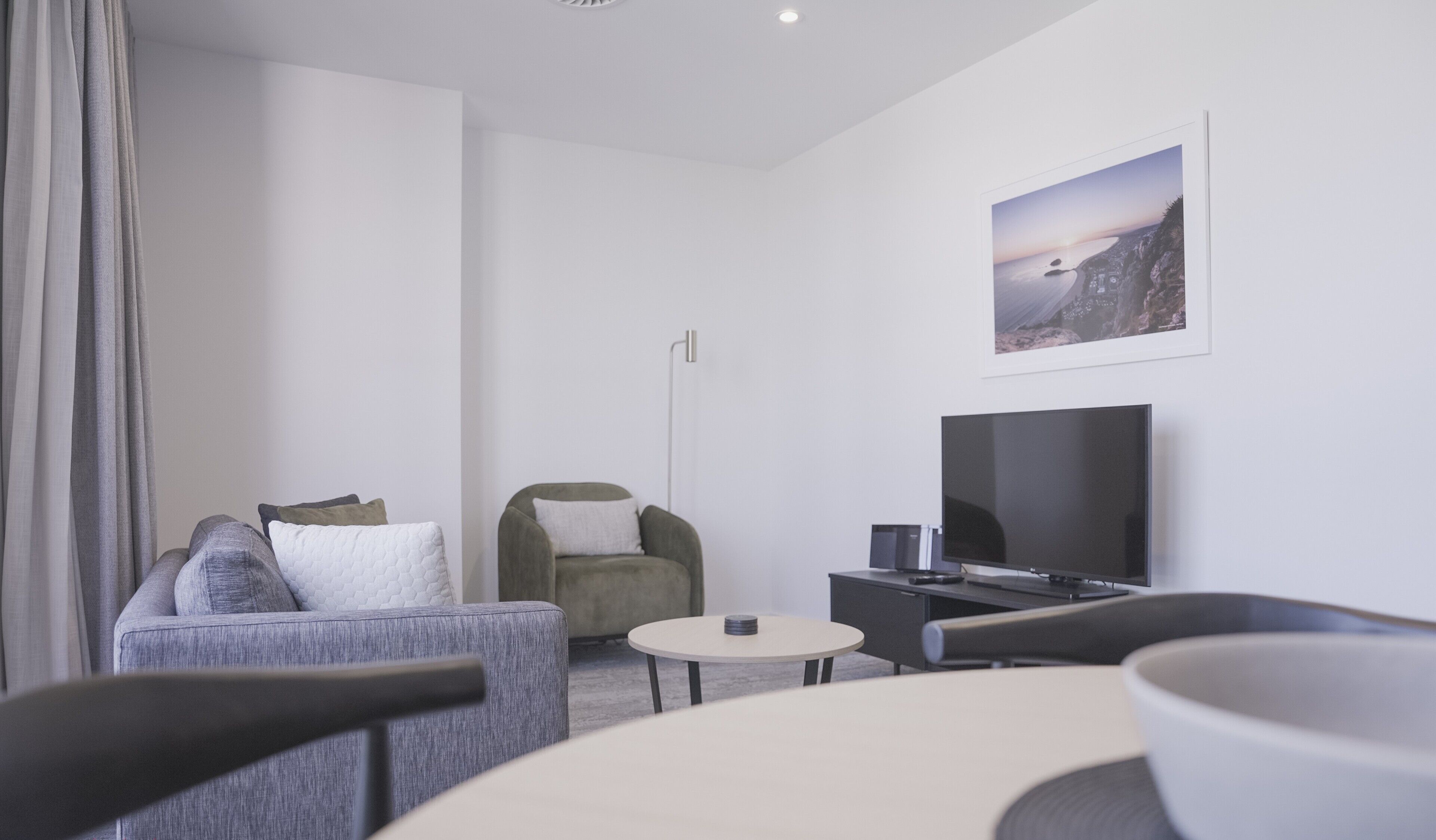 A modern hotel room with a grey sofa, a green armchair, a round coffee table, a flat-screen TV, and a framed picture on the wall.