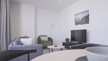 A modern hotel room with a grey sofa, a green armchair, a round coffee table, a flat-screen TV, and a framed picture on the wall.