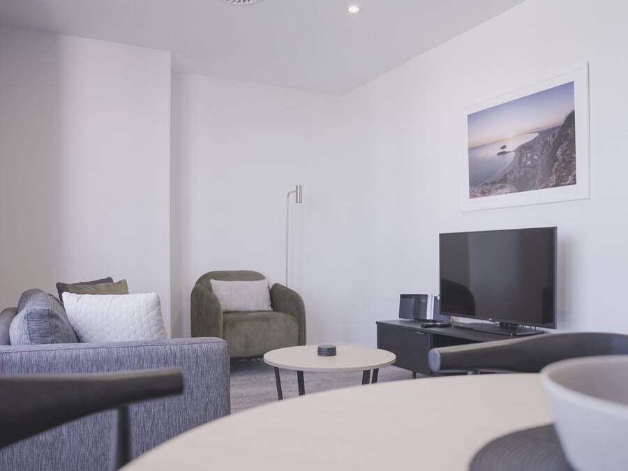 A modern hotel room with a grey sofa, a green armchair, a round coffee table, a flat-screen TV, and a framed picture on the wall.