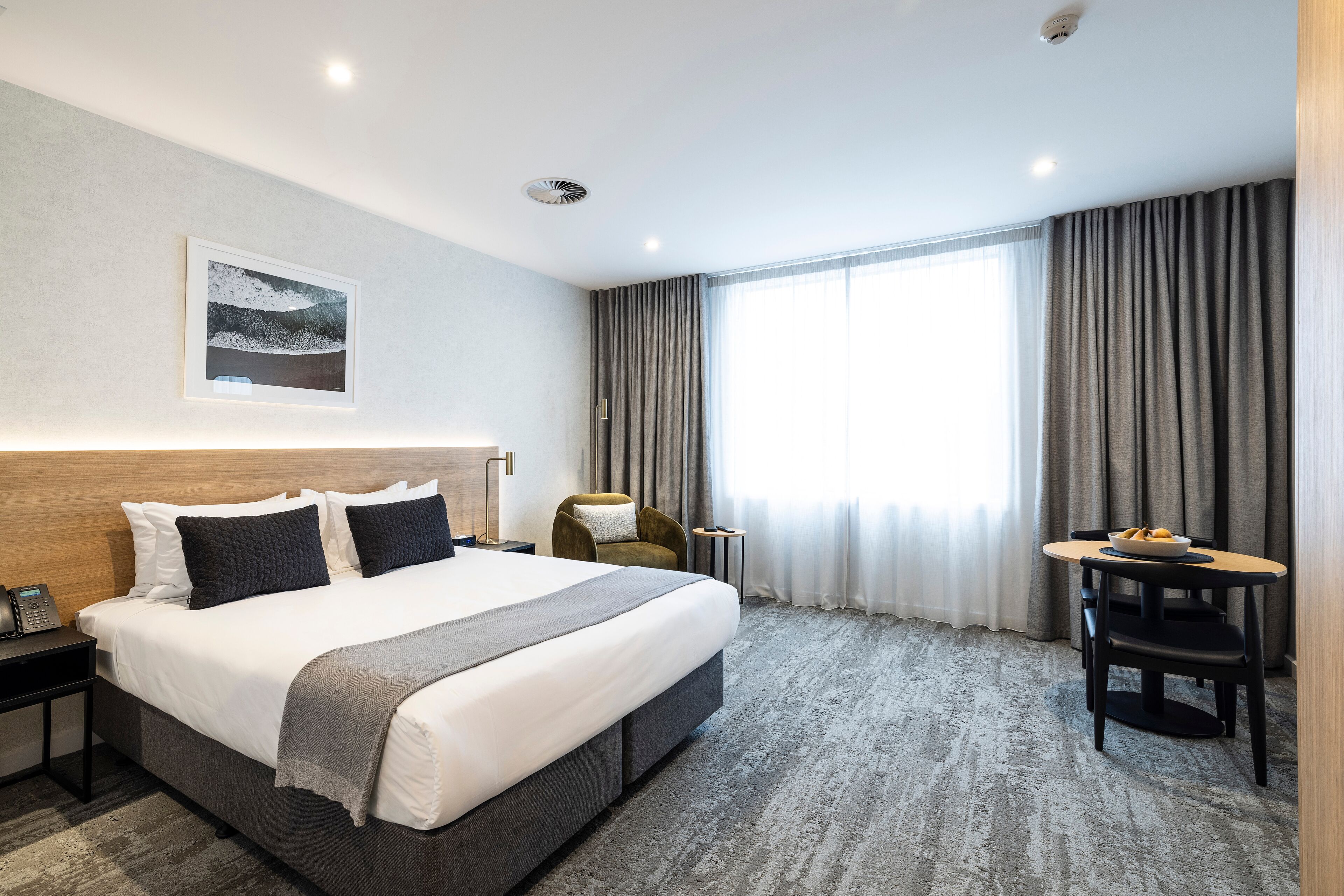 Hotellihuone, jossa on suuri sänky, työpöytä tuolilla, pieni pöytä hedelmäkulhon kanssa ja ikkuna, jossa on verhot.