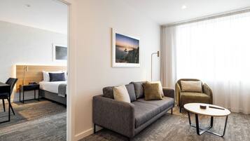 Una habitación de hotel moderna con un sofá gris, una mesa de centro redonda, una cama con cabecera, una mesita de noche con lámpara y un ventanal con cortinas.