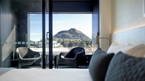 A modern hotel room with a large bed, two chairs, a desk, and a view of a mountain. - Quest Mt Maunganui  (Tauranga)