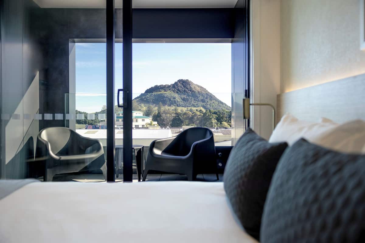 A modern hotel room with a large bed, two chairs, a desk, and a view of a mountain.
