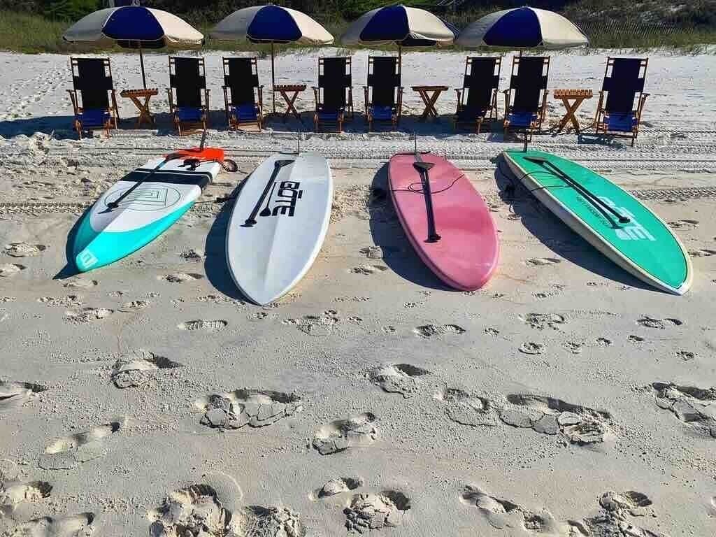Four surfboards lined up on a sandy beach with beach chairs and umbrellas in the background.