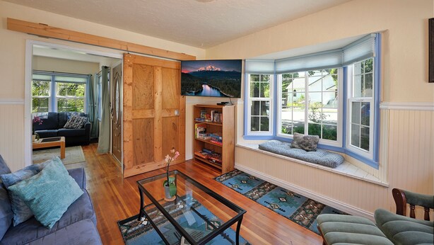 A cozy living room with a wooden door, a bookshelf, a glass coffee table, and a bay window with a view of the outdoors. - Charming 4BR Home in a Great Downtown Location (Mount Shasta)