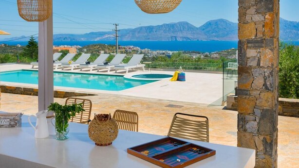 Un espace aménagé au bord de la piscine, avec des chaises longues, une table sur laquelle on peut jouer à des jeux de société, et une vue sur la mer et les montagnes.