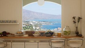 A breakfast table with a scenic view of a coastal town and mountains, a fruit bowl, a coffee pot, and a vase with flowers. - Villa Queen with a heated pool (Elounda)