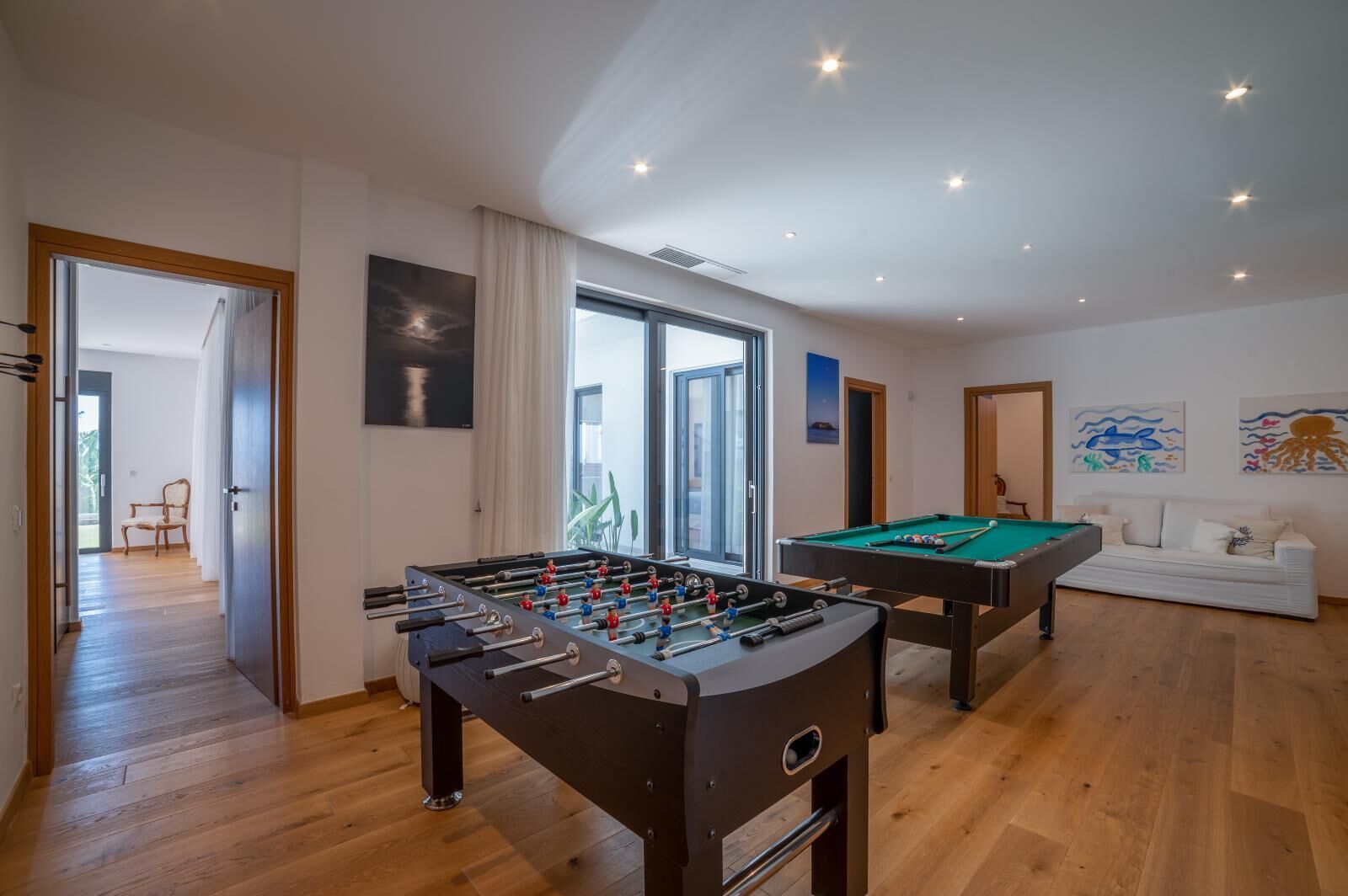 A room with a foosball table, a pool table, and a sofa. There are windows and artwork on the walls.