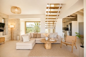 A modern living room with a sectional sofa, a round coffee table, a wicker chair, a staircase, and a kitchen area.