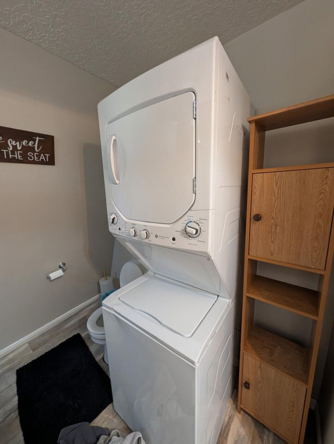 A laundry area with a stacked washer and dryer, a toilet, a black rug, and a wooden shelf unit.