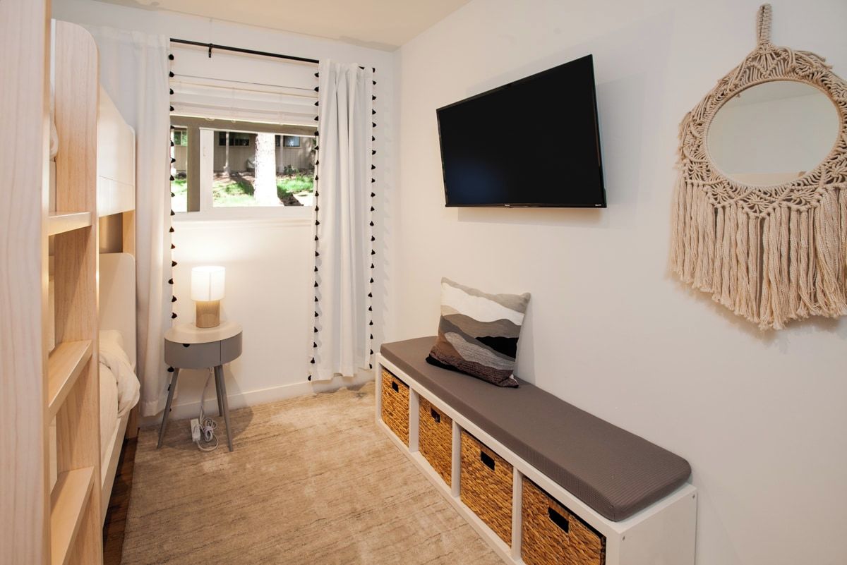 A room with a TV mounted on the wall, a bench with storage underneath, a bedside table with a lamp, and a macrame wall hanging.