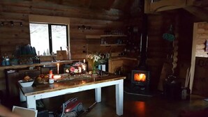 Interior rústico de una cabaña con detalles de madera, una mesa de comedor con velas, un estufa de leña y una ventana con vistas a un bosque.