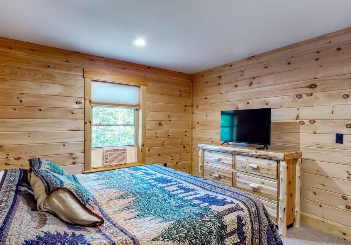 A wooden-paneled bedroom with a bed, a dresser, a television, and a window.