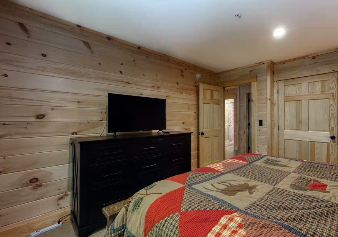 A bedroom with wooden walls, a black dresser, a television on top, and a quilted bedspread.