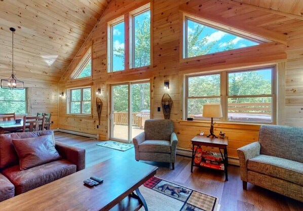 Cozy cabin with a living area, sofa, coffee table, and fireplace, surrounded by large windows and trees.