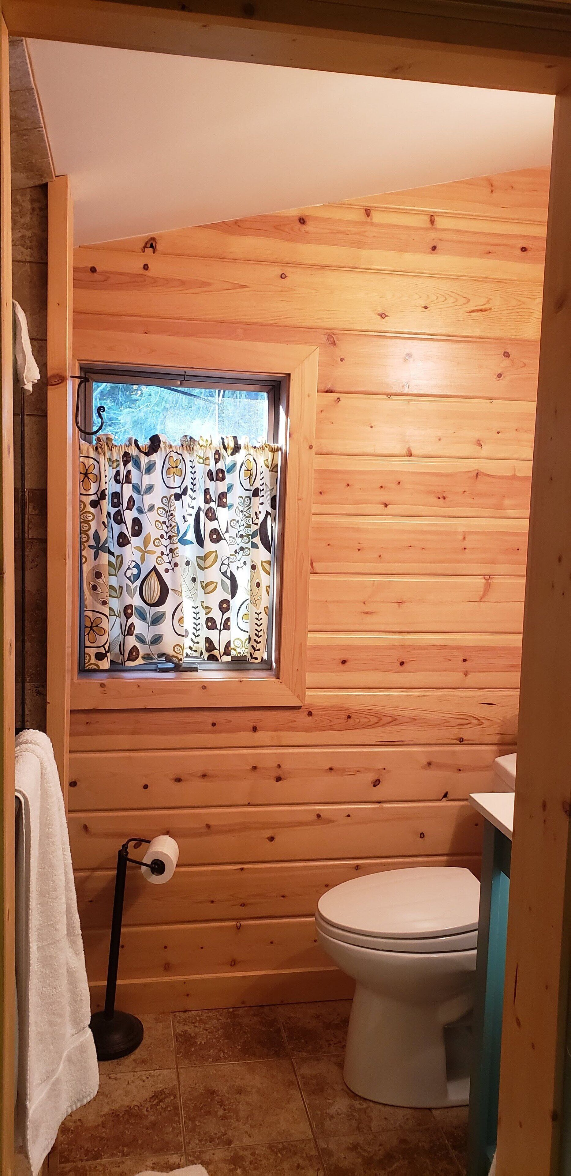 Un baño con paneles de madera, un inodoro, una ventana con cortinas estampadas y una toalla colgada en un perchero.