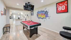A pool table in a room with a kitchen in the background, a TV mounted on the wall, and a red sports jersey with the number 80. - The Ultimate Man Cave Haven: Arcade Room/ Pool Table (Houston)