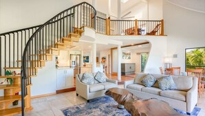 A spacious living area with a curved staircase, a dining area with wooden furniture, and a view of the outdoors. - Luxury 2-Story Oceanfront Condo w/ Views & Pool (Wailua)