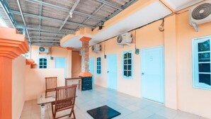 Amplia habitación con piso de baldosas, una mesa de vidrio, dos sillas de madera, un tapete negro y un techo con vigas a la vista y unidades de aire acondicionado.