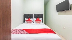 Una habitación pequeña con cama individual, puerta de madera, televisor de pantalla plana en la pared y colcha de cama roja y blanca.