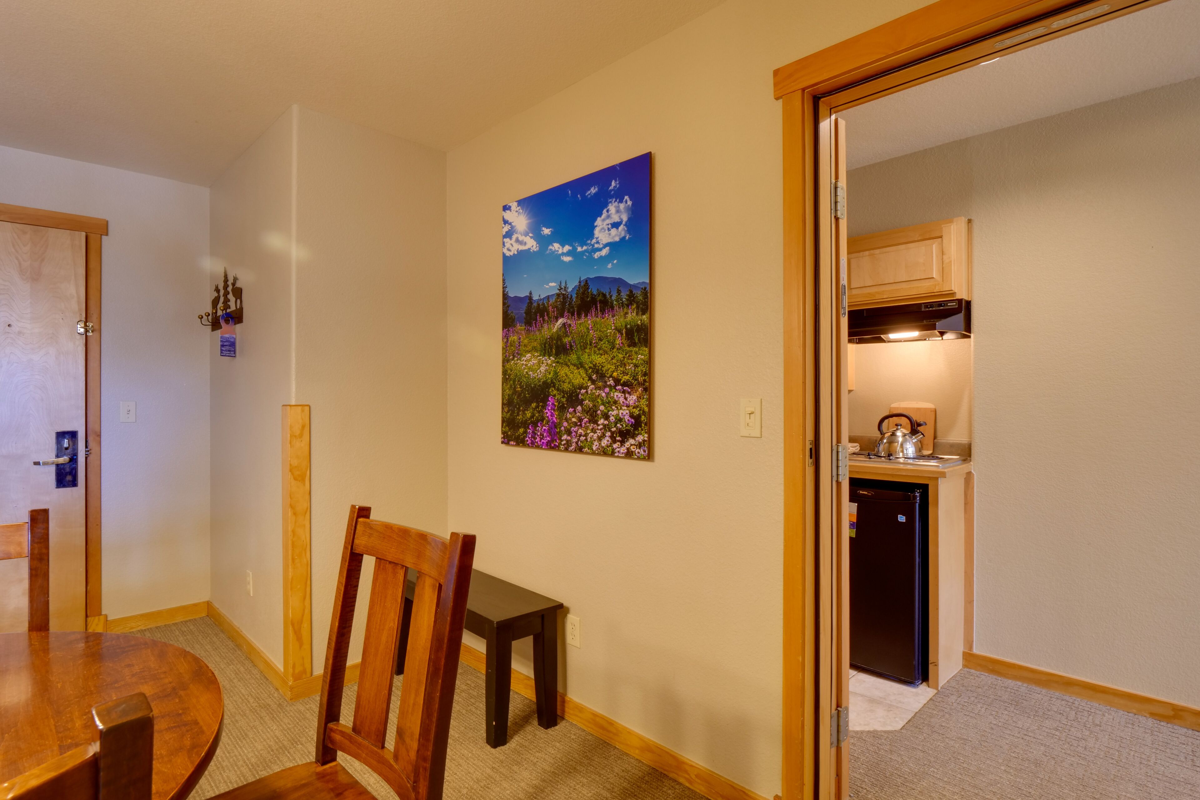 Un coin repas avec une table et des chaises, un tableau représentant un paysage et une cuisine visible à travers une porte ouverte.