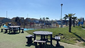 Explore the playground area with swings, climbing frames, picnic tables, and benches amidst lush greenery and palm trees.
