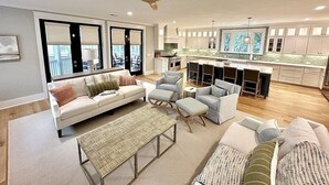 Amplia sala de estar con un sofá, otomanas y una mesa de centro. Se ve una cocina con armarios blancos y un comedor al fondo.