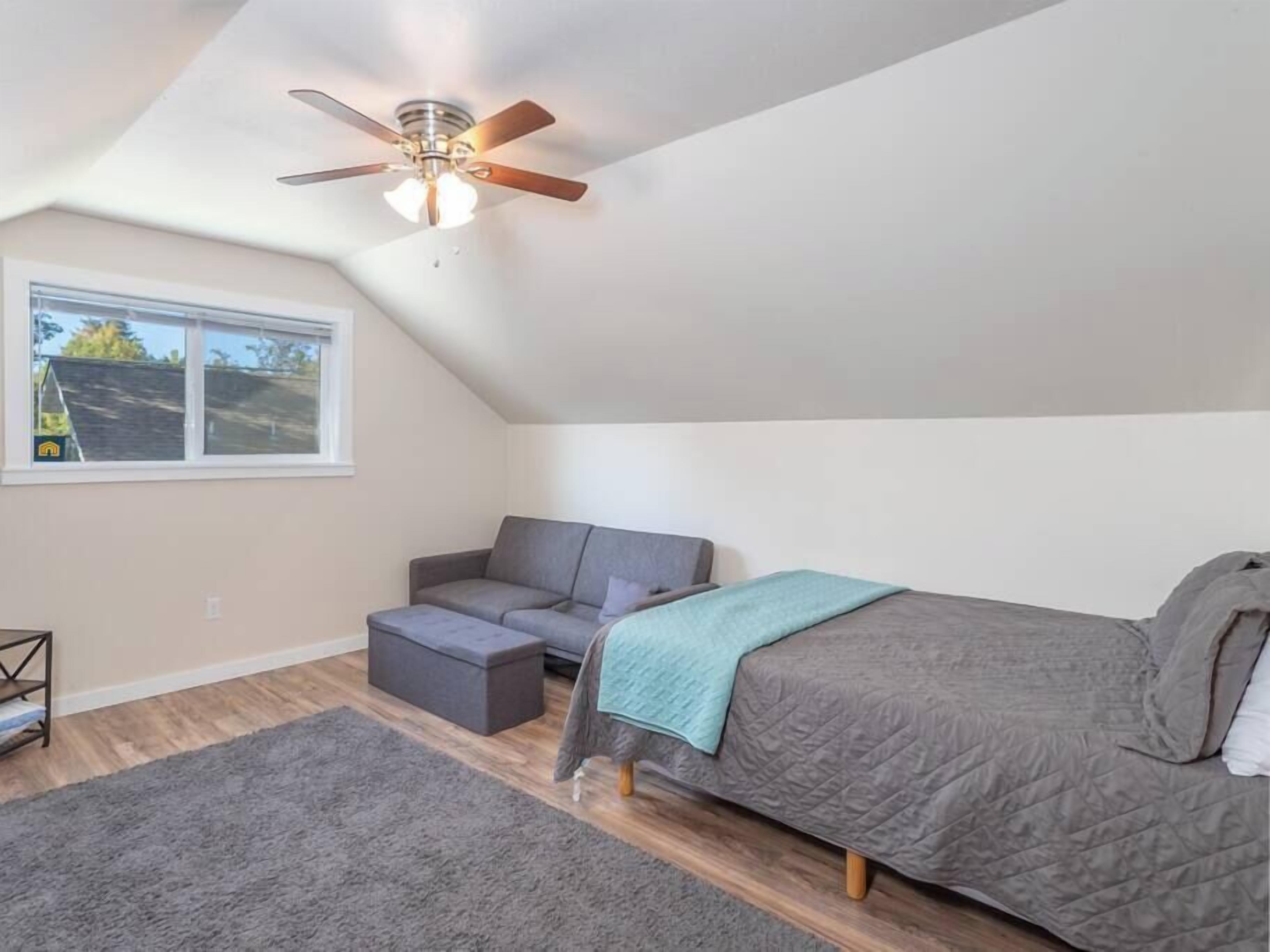 A bedroom with a sloped ceiling, a ceiling fan, a bed, a gray sofa, a small ottoman, and a window with a view of a house.