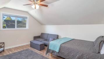 A bedroom with a sloped ceiling, a ceiling fan, a bed, a gray sofa, a small ottoman, and a window with a view of a house.