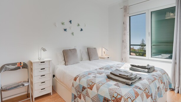 A bedroom with a bed, a nightstand, a dresser, a chair, and a window with a view. - Alta Garajau II (Santa Cruz)