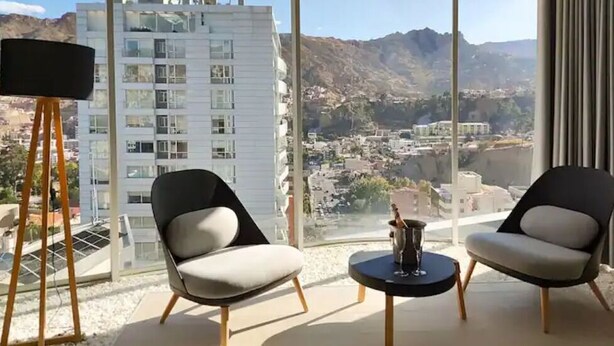 A modern room with two chairs and a small table, large windows with a mountain view, and a floor lamp. - Beautiful Penthouse in the best area of the LP city (Zona Calacoto La Paz)