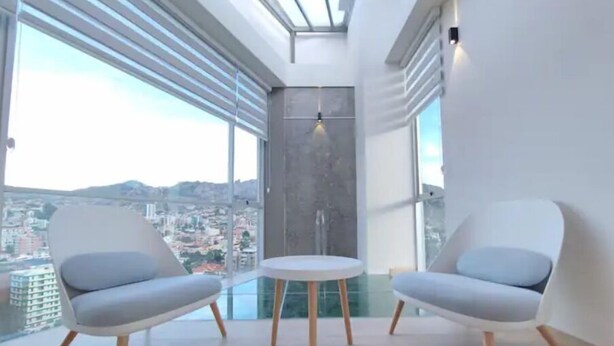 A modern room with two grey chairs and a small table, large windows with blinds, and a city view. - Beautiful Penthouse in the Best Area of the City (Zona Calacoto La Paz)